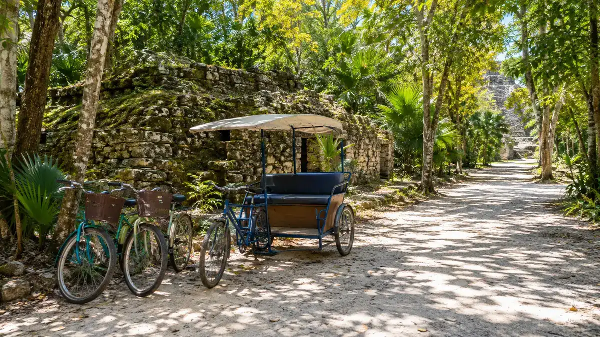 Two rental bicycles and a pedal tricycle taxi parked on a shaded white path beside a mossy ruin at Coba