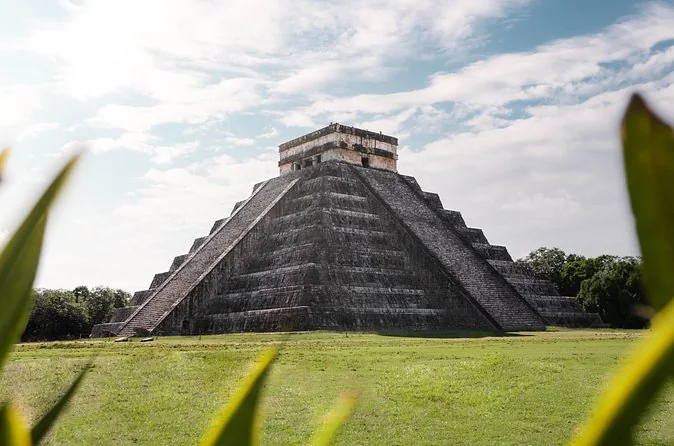 The stepped El Castillo pyramid at Chichen Itza on a wide lawn, framed by leaves