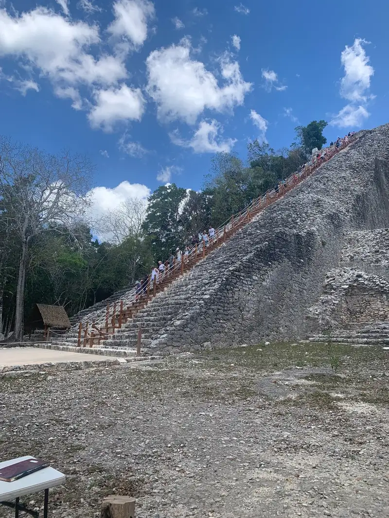 Visitors climbing the wooden staircase with orange handrails up the grey stone face of Nohoch Mul