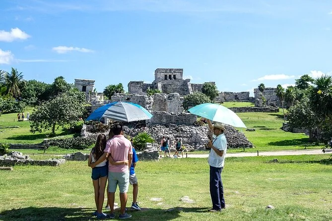 Visitors with umbrellas and a guide on the lawn at Tulum ruins, temples behind