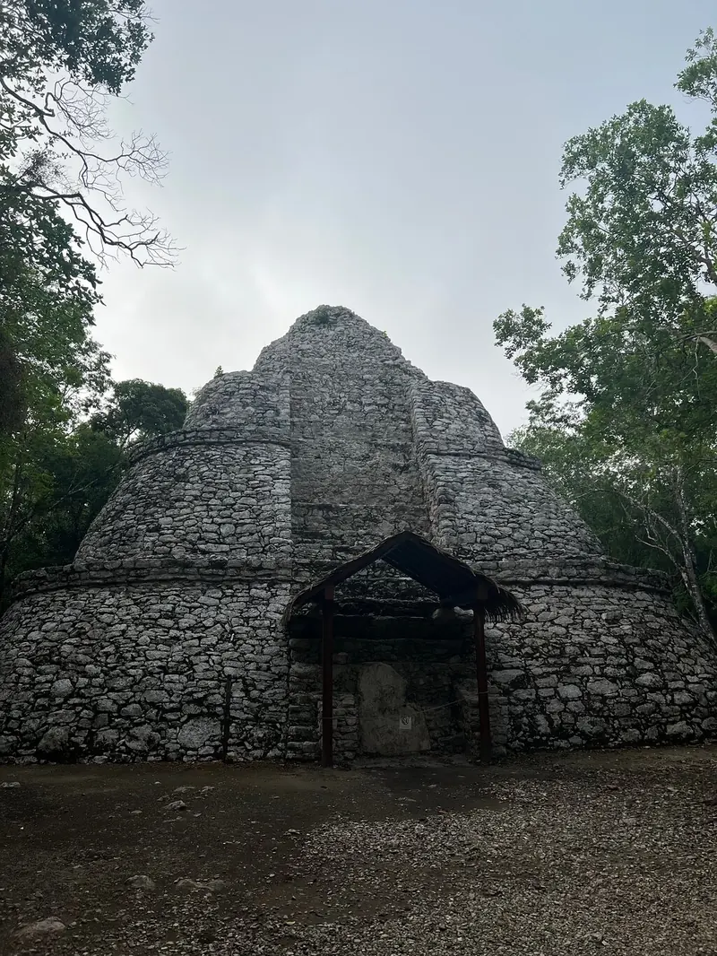 A tall stepped structure with rounded corners and a small doorway at its base, among trees